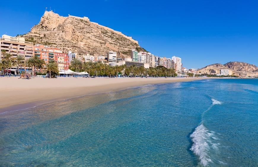 Playa del postiguet en Alicante con el castillo al fondo, arena y aguas cristalinas. Cielo azul ideal para una pedida de mano y entregar anillo de compromiso
